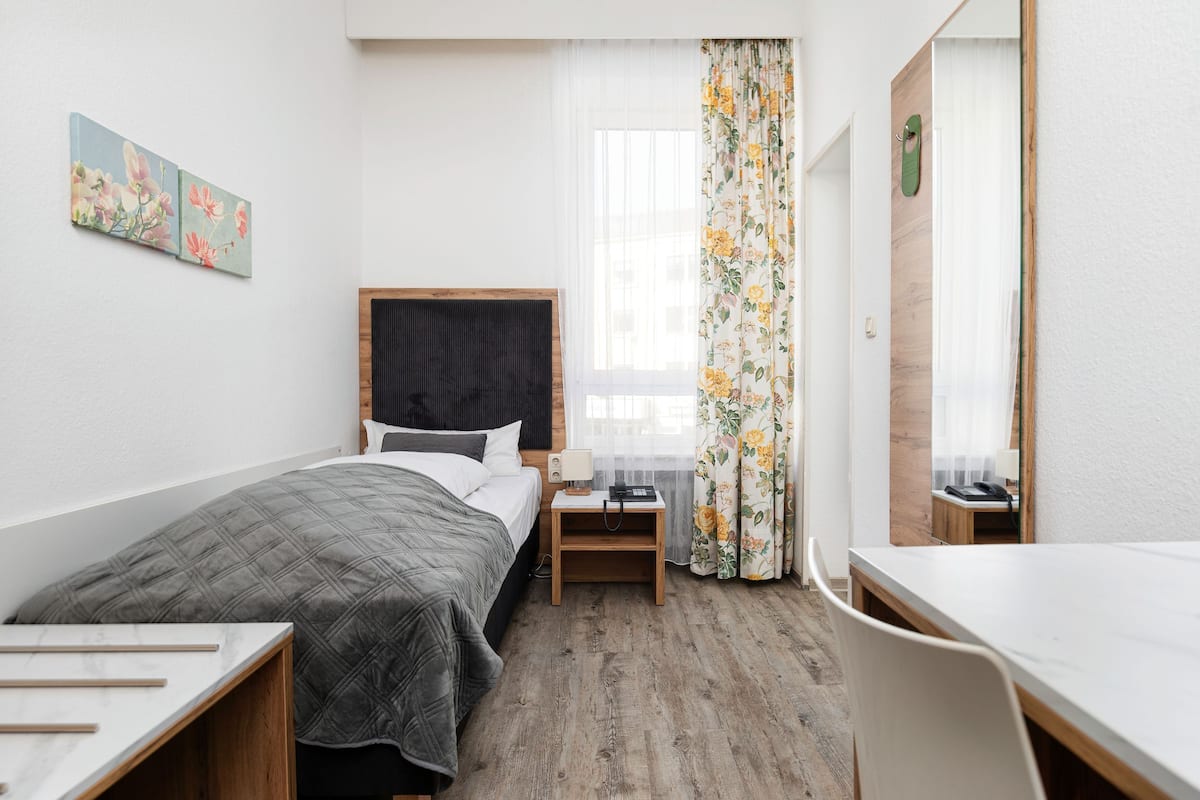 A compact hotel room with a bed, bedside table, chair, and a small desk.