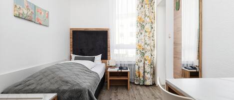 A compact hotel room with a bed, bedside table, chair, and a small desk.