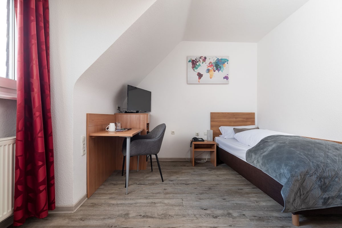 A hotel room with a bed, a desk with a chair, a television, a small bedside table, and a world map artwork on the wall.