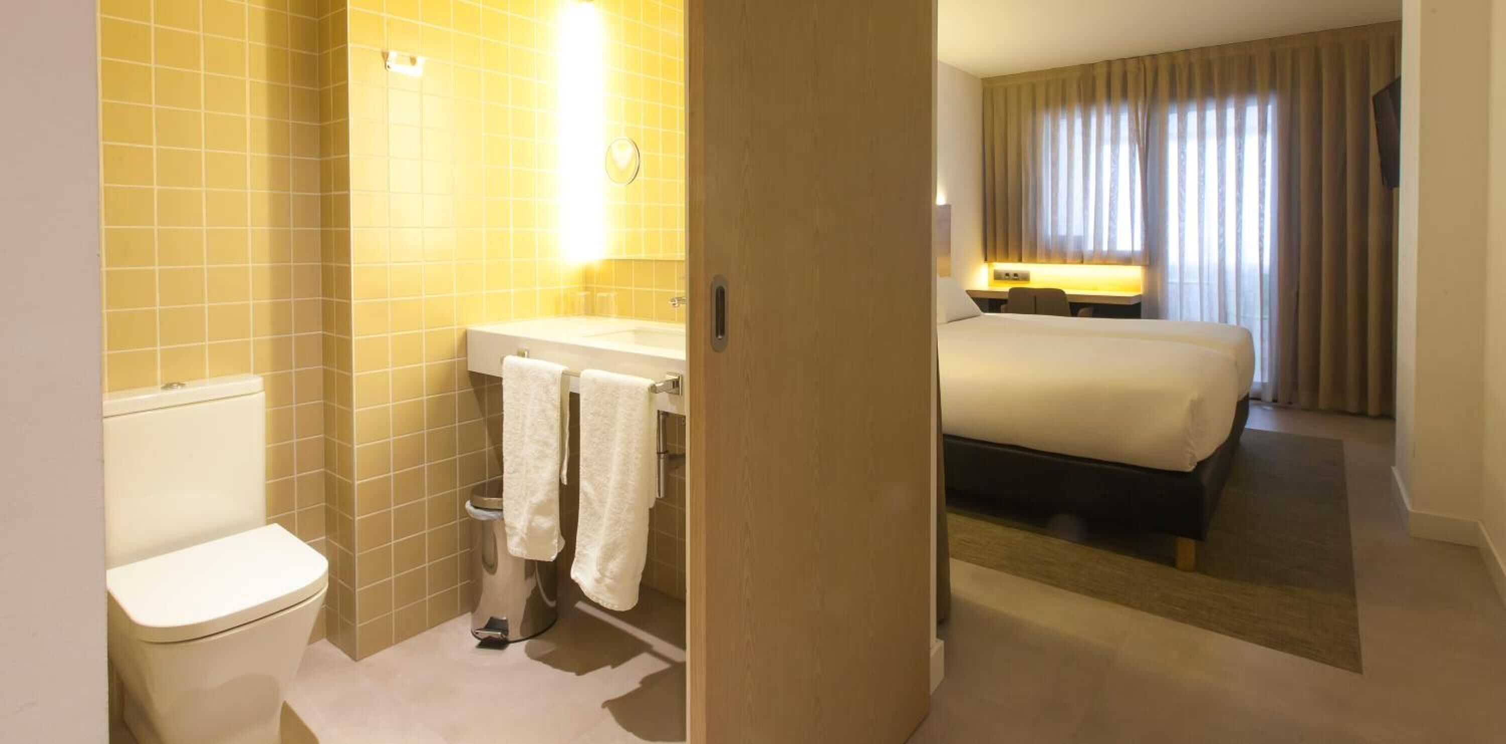 Ein Badezimmer mit Toilette, Waschbecken und Handtuchhalter. Das Zimmer verfügt außerdem über ein Bett mit Nachttisch und ein Fenster mit Vorhängen.