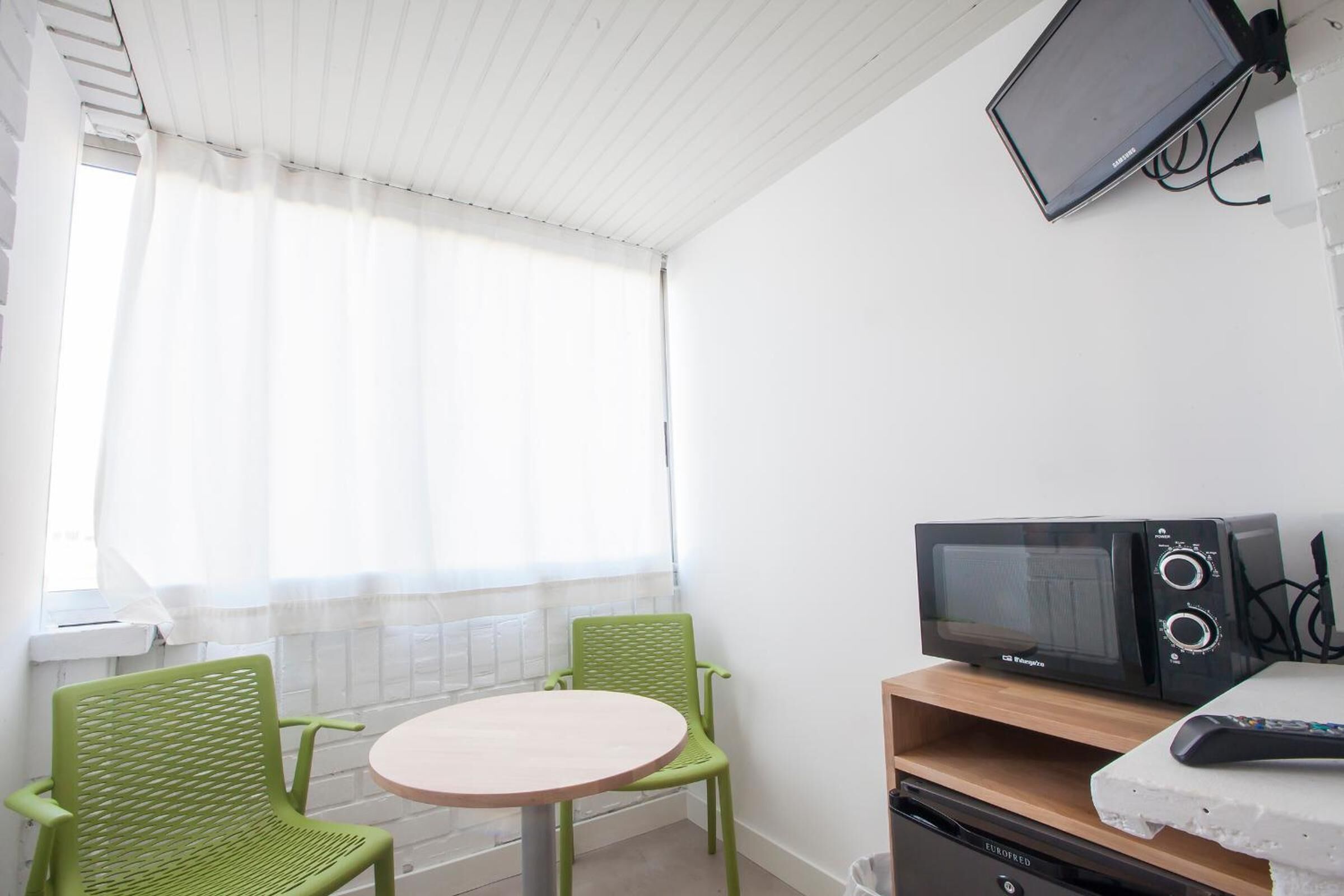 A small room with a round table and two green chairs, a microwave, and a television on a wooden shelf.