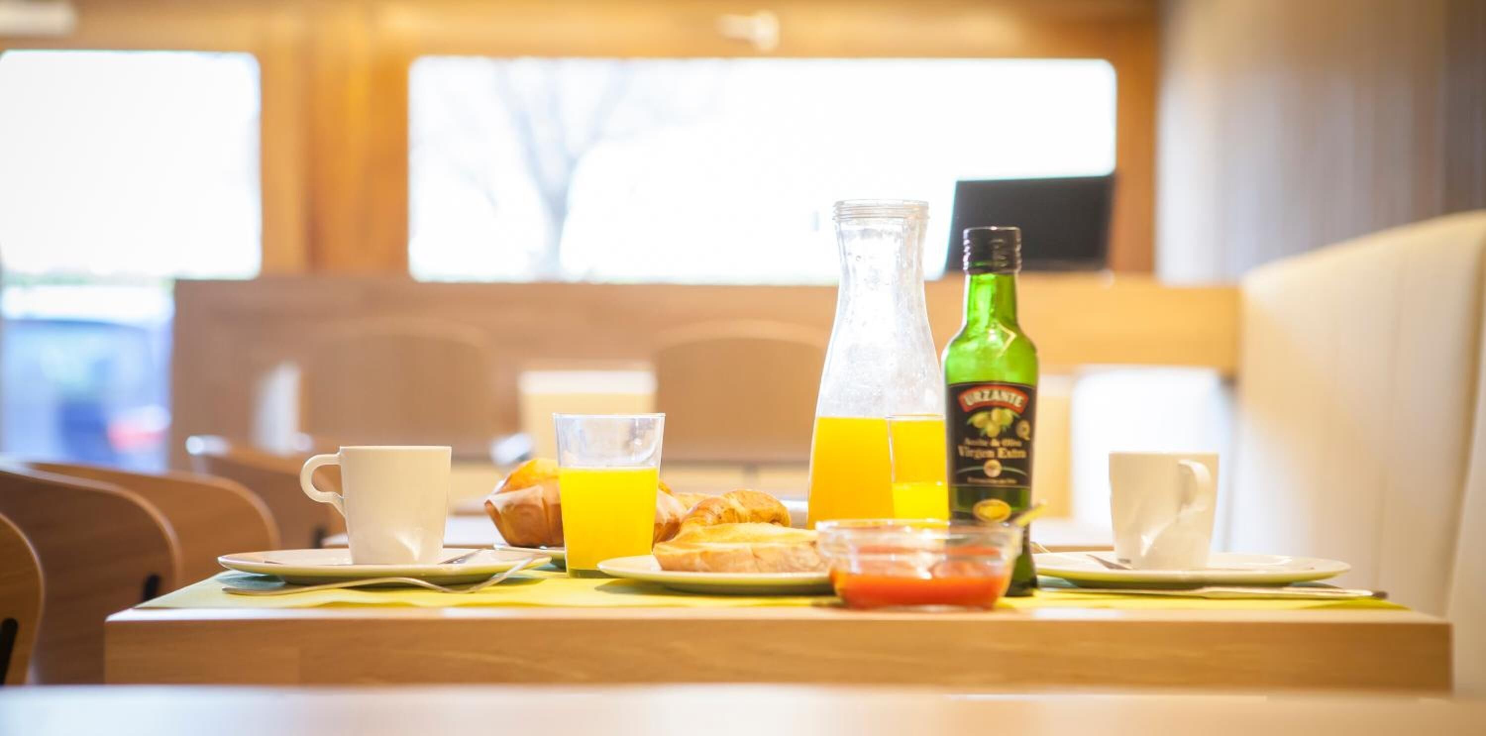 Un plateau de petit-déjeuner avec des croissants, du jus d’orange et une bouteille de jus biologique, disposé sur une table en bois dans une pièce chaleureuse avec vue sur l’extérieur par la fenêtre.