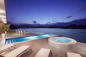 Uma área de piscina com hidromassagem e espreguiçadeiras com vista para um corpo de água ao entardecer.