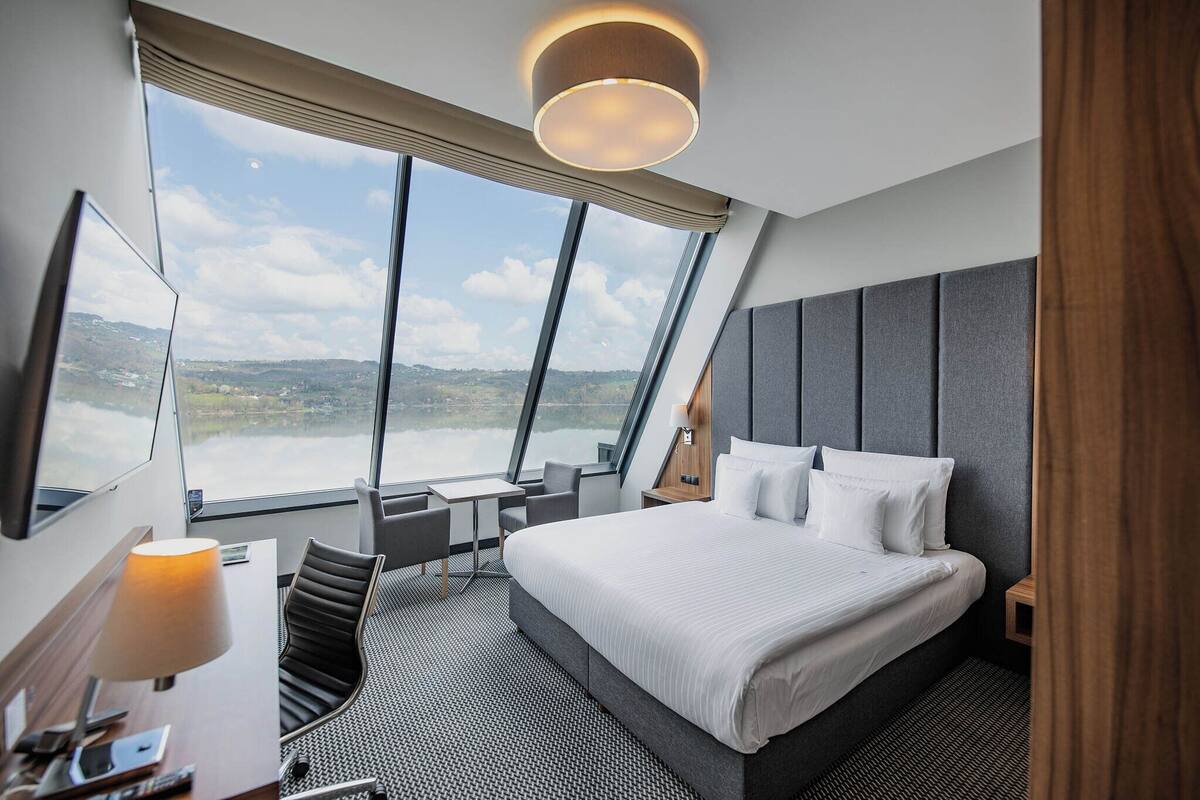 A modern hotel room with a large bed, a desk, a chair, and a view of the outdoors.