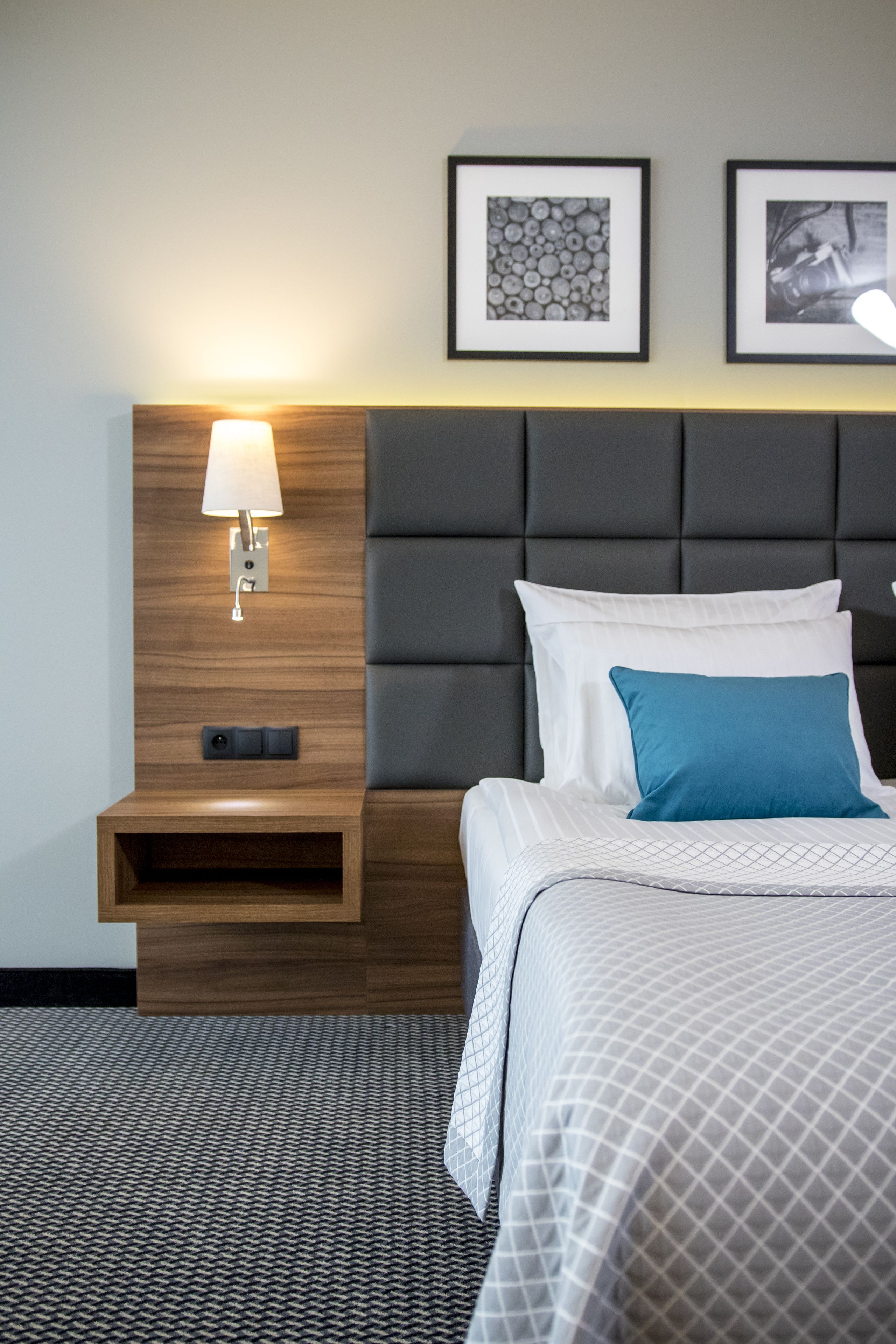 A hotel room with a bed, a nightstand, a wall-mounted lamp, and two framed pictures.