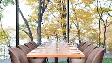 A dining table set with chairs, a vase with flowers, and a view of autumn trees through large windows.