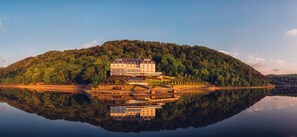 A large, multi-story building with a reflection in the water, surrounded by a forested hill.