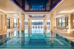 A large indoor swimming pool with clear blue water, surrounded by lounge chairs and large windows offering views of the landscape. - Heron Live Hotel (Grodek Nad Dunajcem)