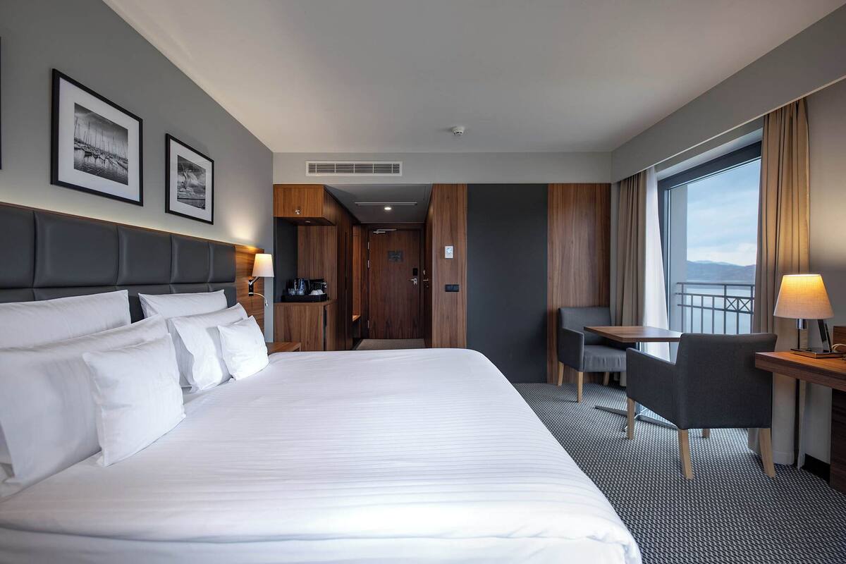 A hotel room with a large bed, a desk with a chair, a view of the sea, and framed pictures on the wall.