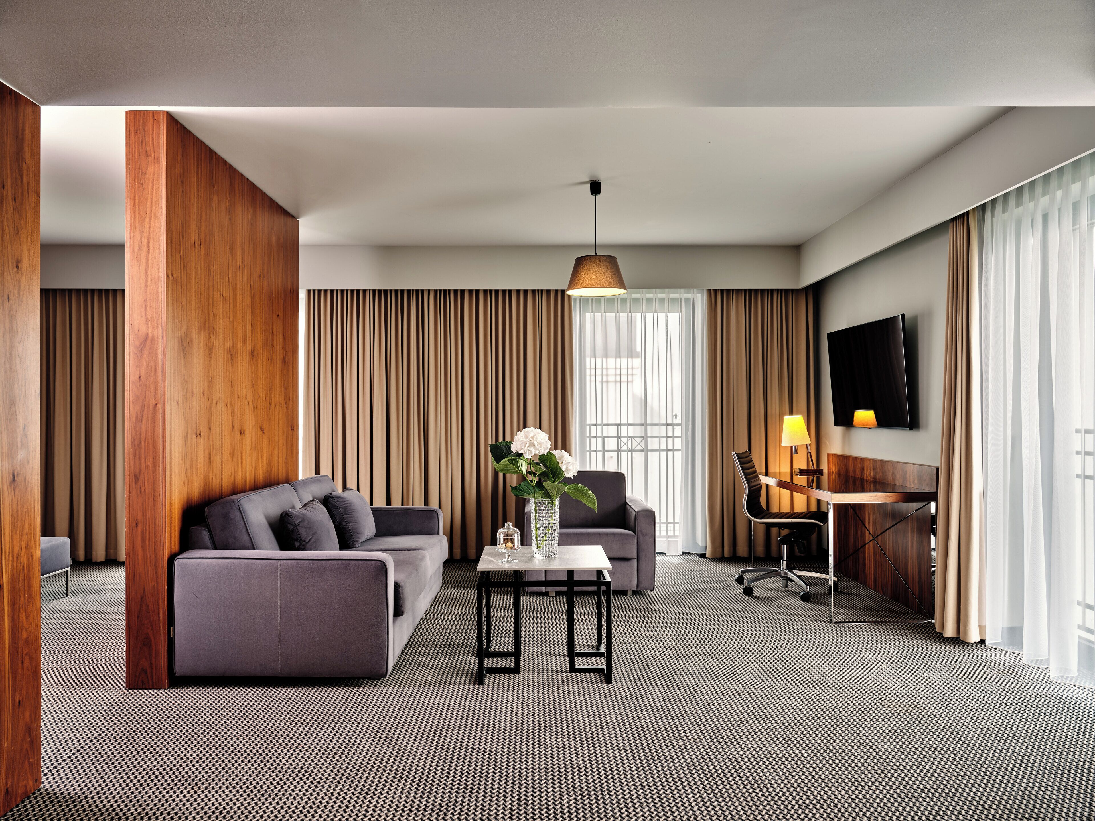 Ein modernes Hotelzimmer mit grauem Sofa, einem kleinen Tisch mit Blumenvase, einem Schreibtisch mit Lampe und einem an der Wand montierten Fernseher.