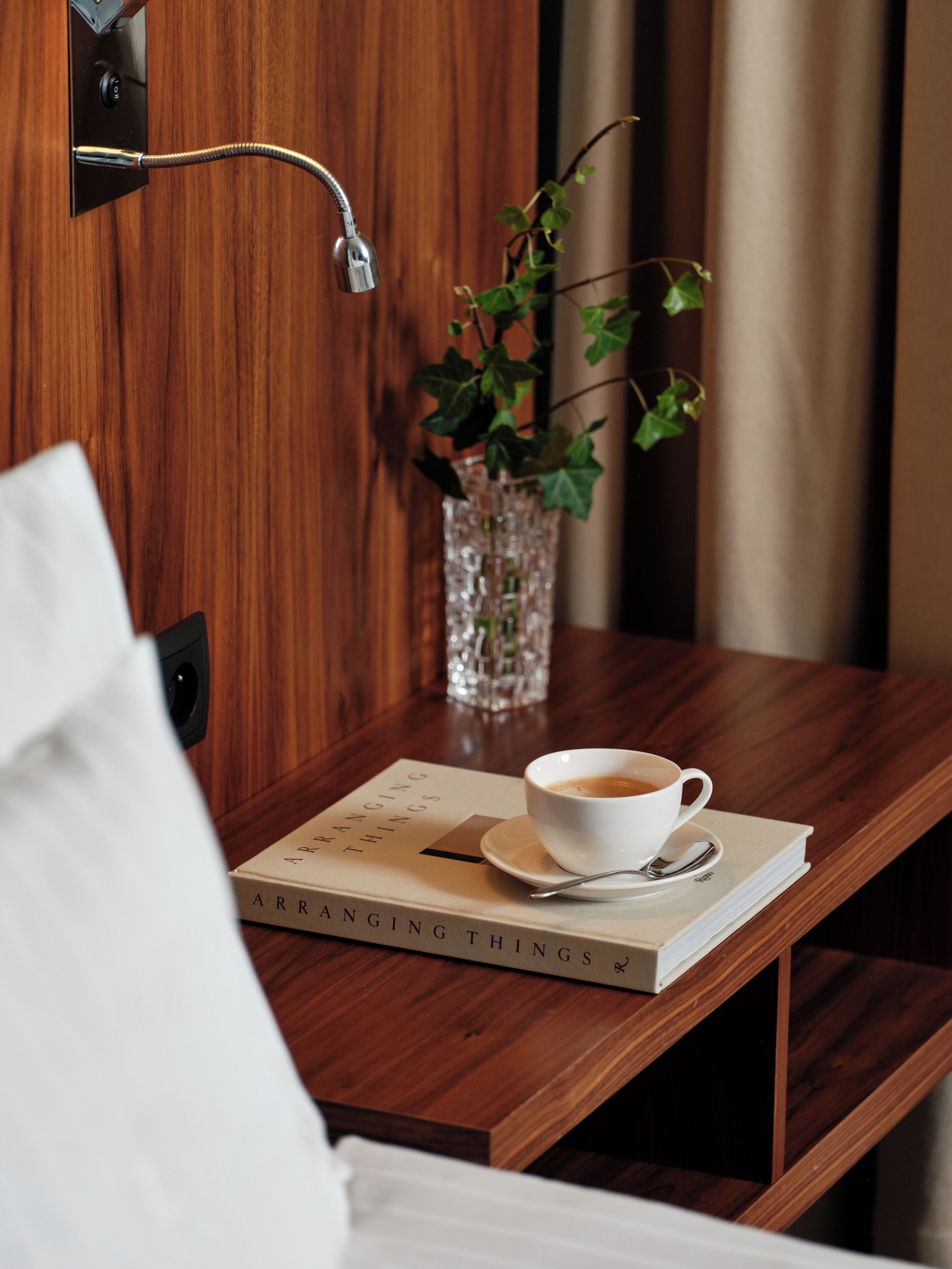 A wooden bedside table with a cup of coffee, a book titled "Arranging Things," and a glass vase with a plant.