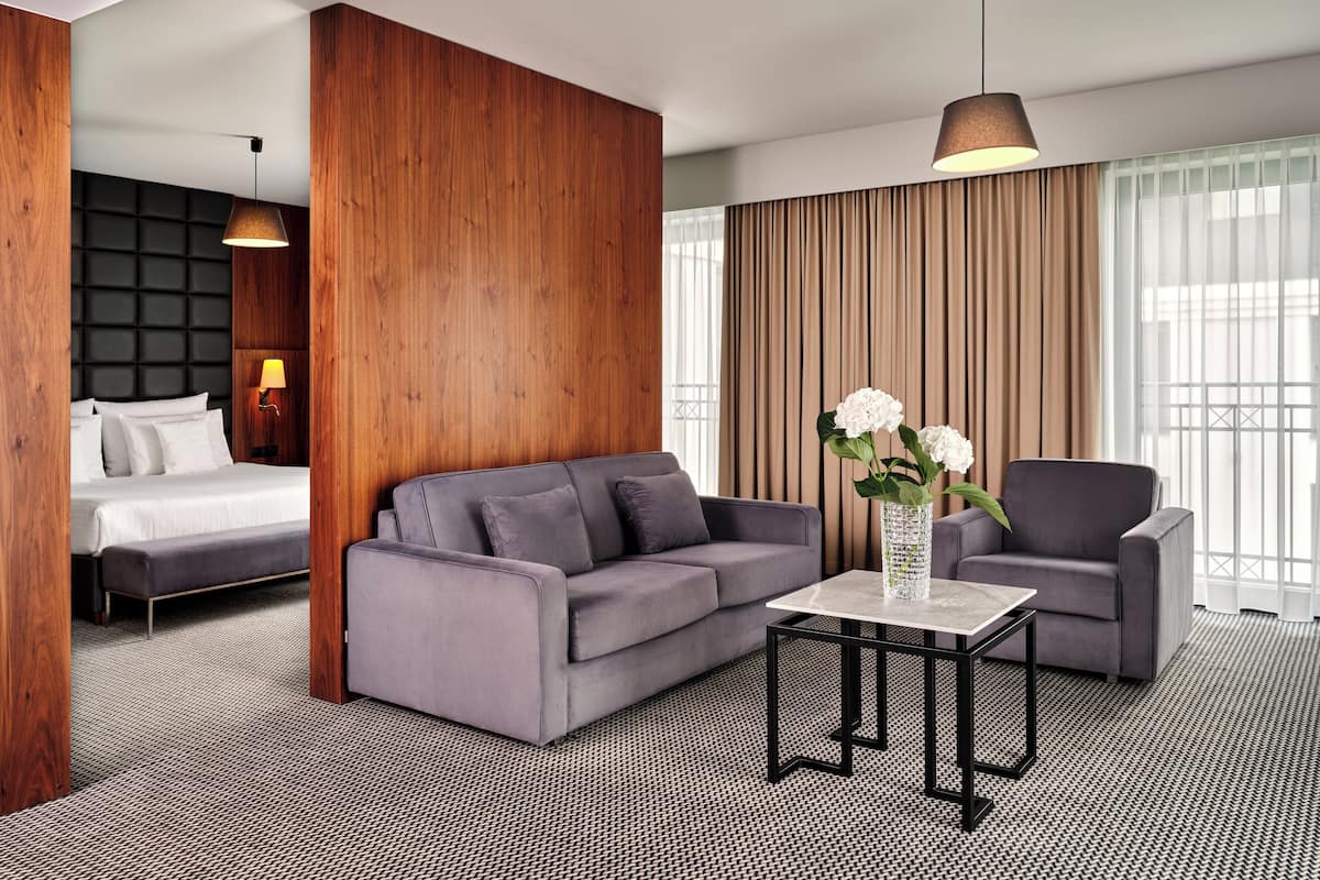 A modern hotel room with a bed, grey sofa, armchair, and a small table with a vase of flowers.