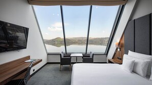 Una habitación de hotel con un ventanal grande, una cama, un escritorio, una silla y un televisor.