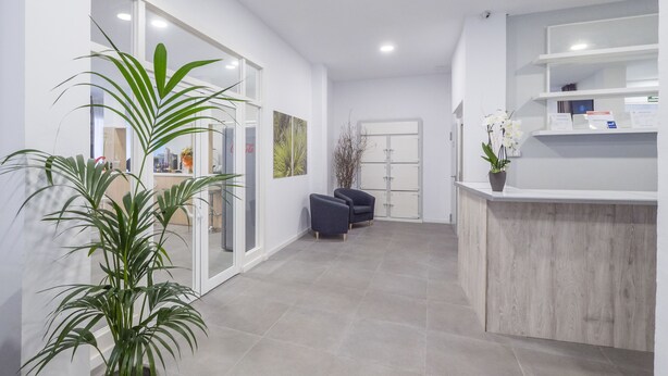 A modern reception area with a wooden reception desk, a seating area with chairs, and potted plants. - Hotel Victoria (Lloret de Mar)