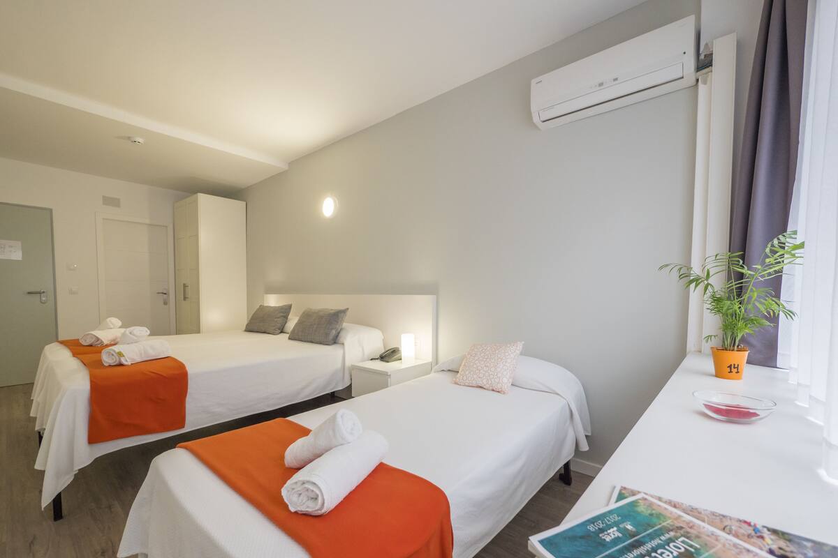 Une chambre d’hôtel avec deux lits, un bureau avec un magazine, un climatiseur et une plante en pot.