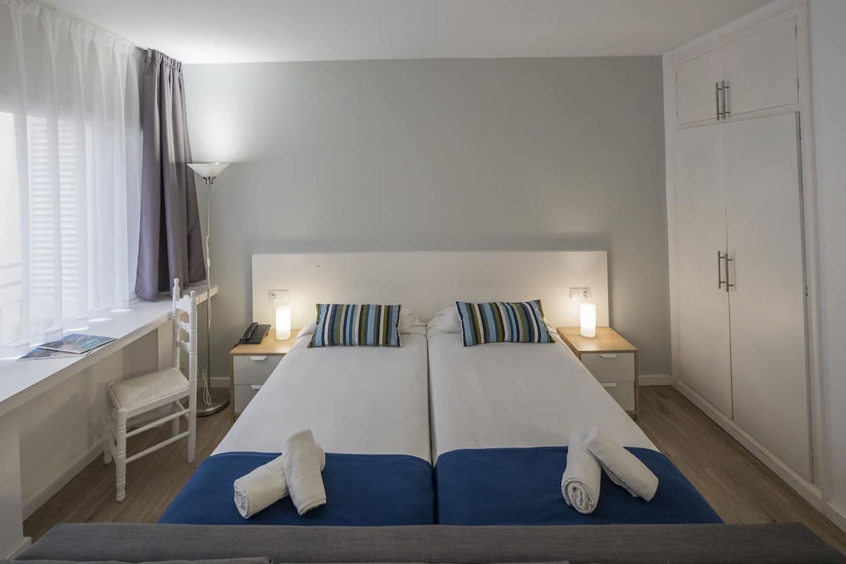 Une chambre d’hôtel avec deux lits, un bureau, une chaise, une armoire et une fenêtre avec des rideaux.