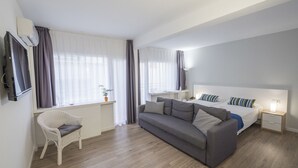 A modern hotel room with a grey sofa, a white chair, a bed with striped pillows, a bedside table, and a TV. - Hotel Victoria (Lloret de Mar)