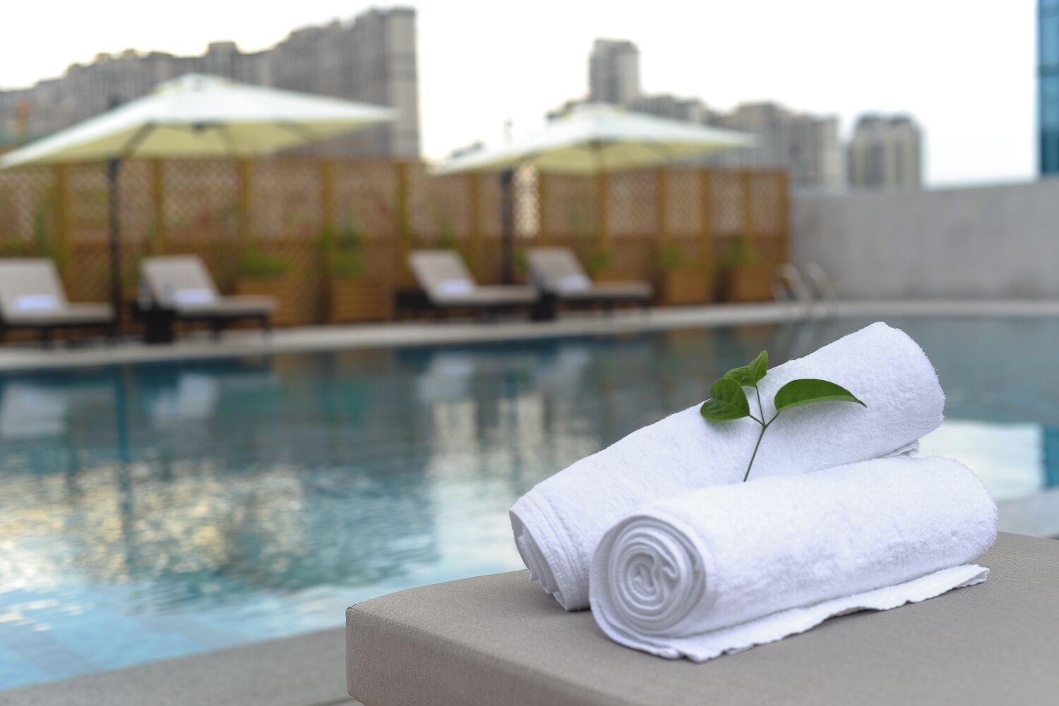 A poolside area with lounge chairs and umbrellas, a pool, and rolled white towels on a surface.