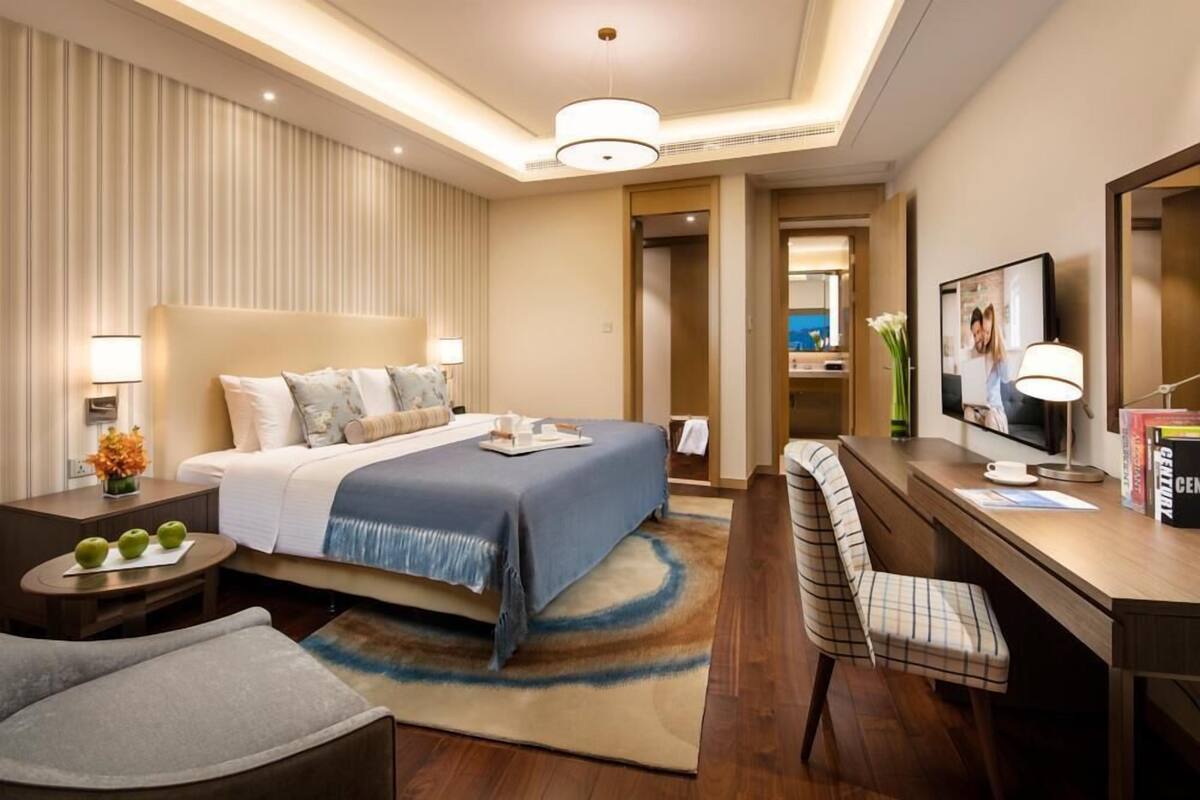 Habitación de hotel con cama, mesitas de noche, escritorio, silla, televisor y baño a la vista a través de una puerta abierta.