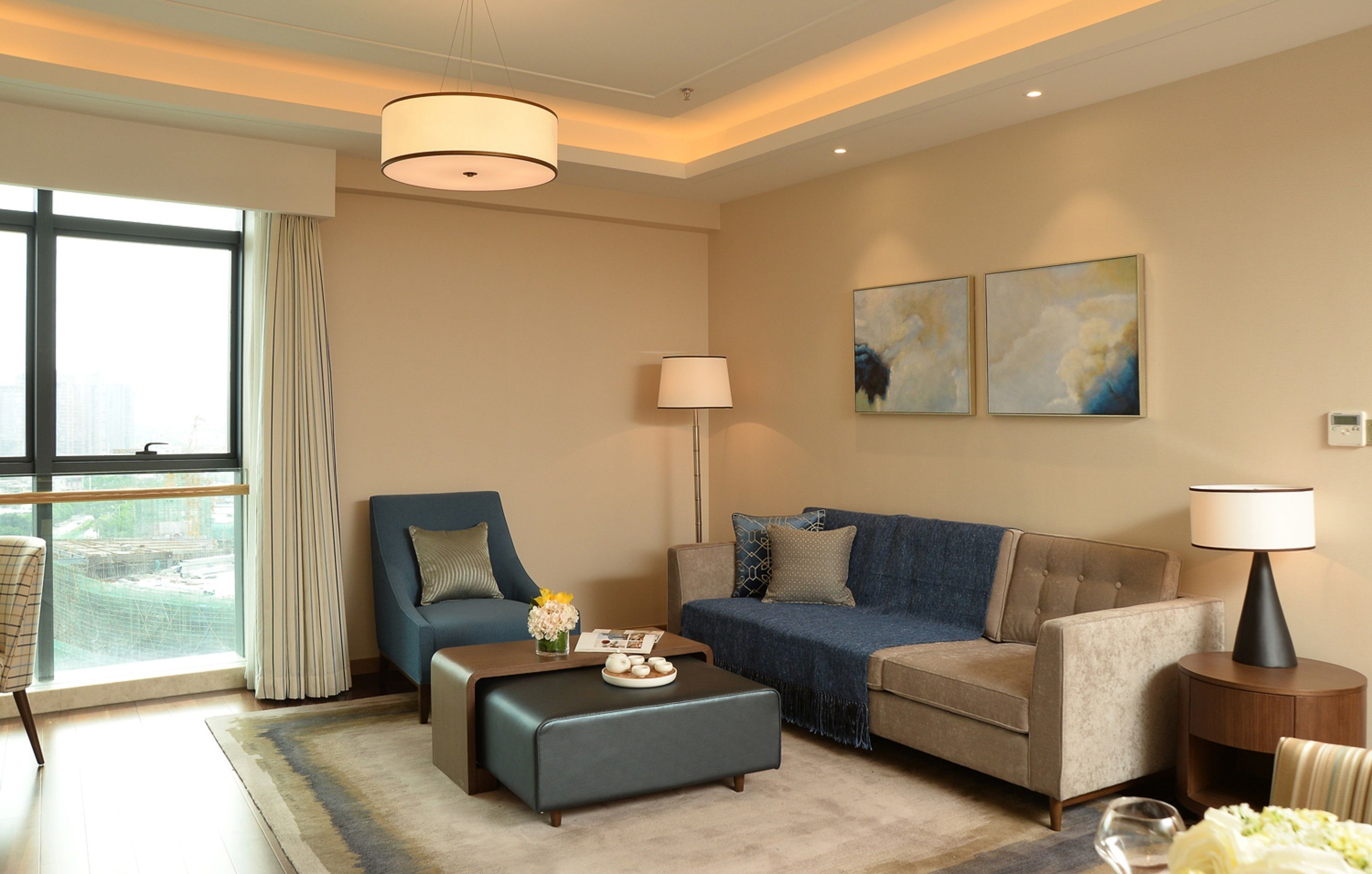 Ein modernes Wohnzimmer mit Sofa, Sessel, Hocker und einem Beistelltisch mit Lampe. Ein großes Fenster mit Vorhängen sowie zwei abstrakte Bilder an der Wand.