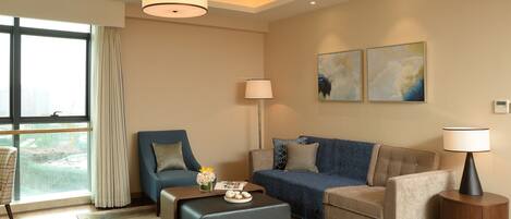 Ein modernes Wohnzimmer mit Sofa, Sessel, Hocker und einem Beistelltisch mit Lampe. Ein großes Fenster mit Vorhängen sowie zwei abstrakte Bilder an der Wand.