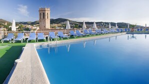 A pool with blue and white striped lounge chairs, a tower, and a view of a hillside town. - Hotel Mercè (Pineda de Mar)