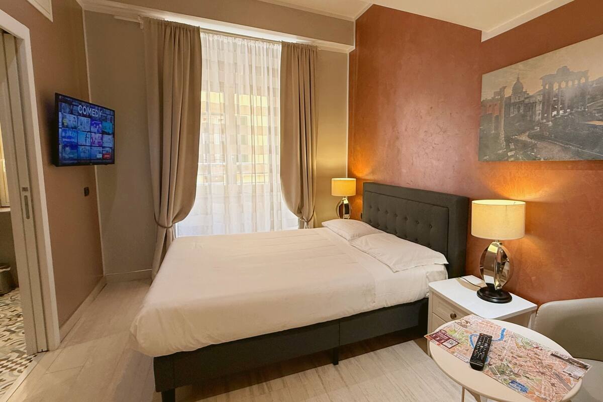 Une chambre d’hôtel avec un lit, deux lampes de chevet, un téléviseur fixé au mur, une carte sur la table et un tableau au mur.