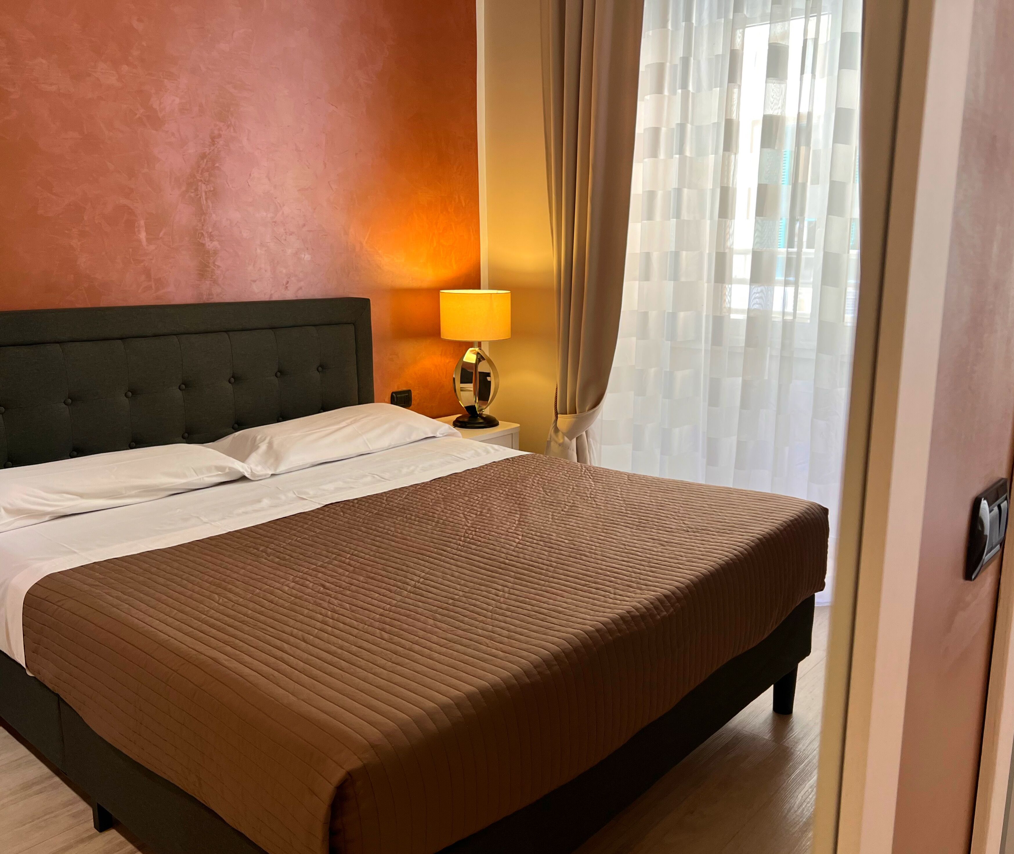 A neatly made bed with a brown bedspread, a bedside lamp, and a window with sheer curtains.