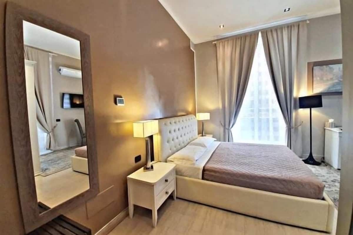 Une chambre d’hôtel avec un grand lit, une table de chevet avec une lampe, un miroir et une fenêtre avec des rideaux.