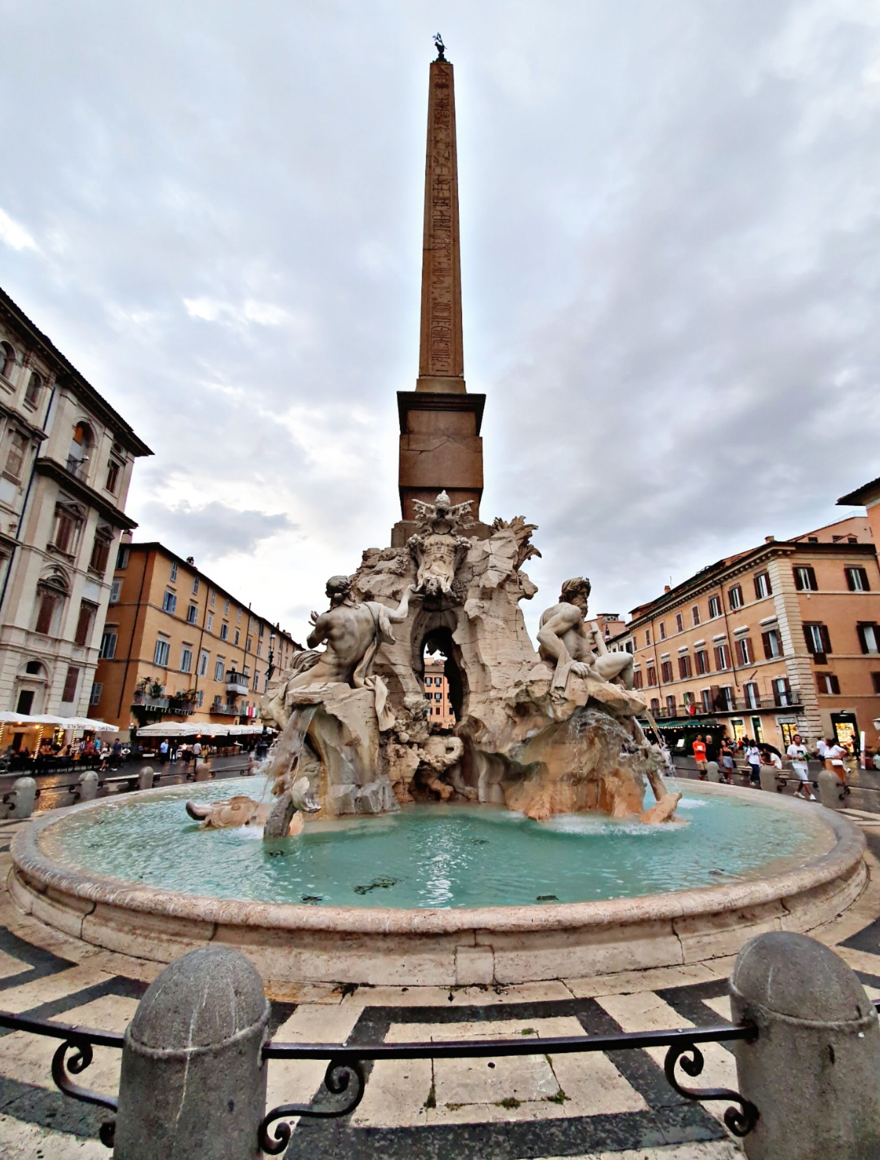 A historic fountain with a tall obelisk and sculptures, surrounded by buildings and a paved area.