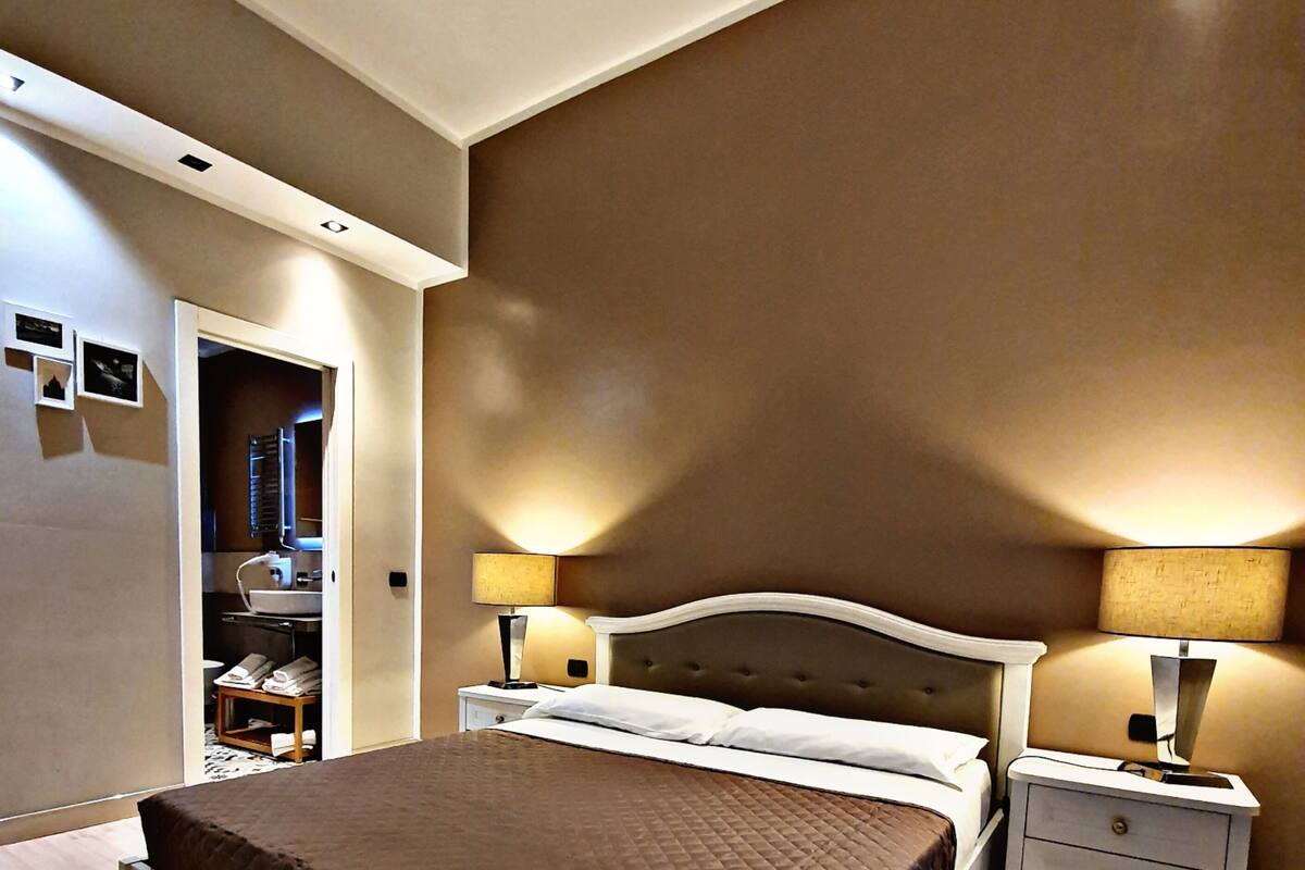 Une chambre à coucher avec un lit, des tables de chevet, une lampe et une salle de bain visible à travers une porte ouverte.