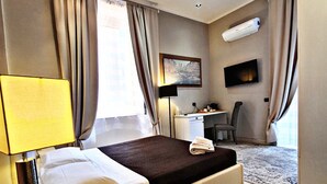A hotel room with a bed, bedside table, lamp, desk, chair, TV, and an air conditioning unit. - Fabio Massimo Guest House (Rome)