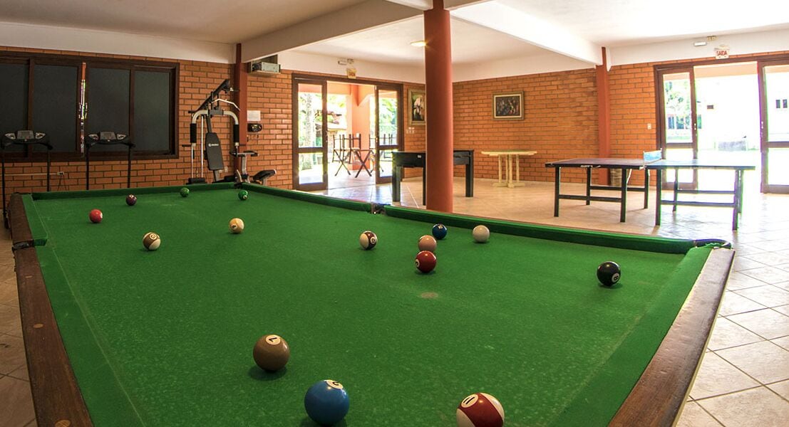 Une table de billard avec des billes, une queue de billard et une table de ping-pong, dans une pièce aux murs en briques et au sol carrelé.
