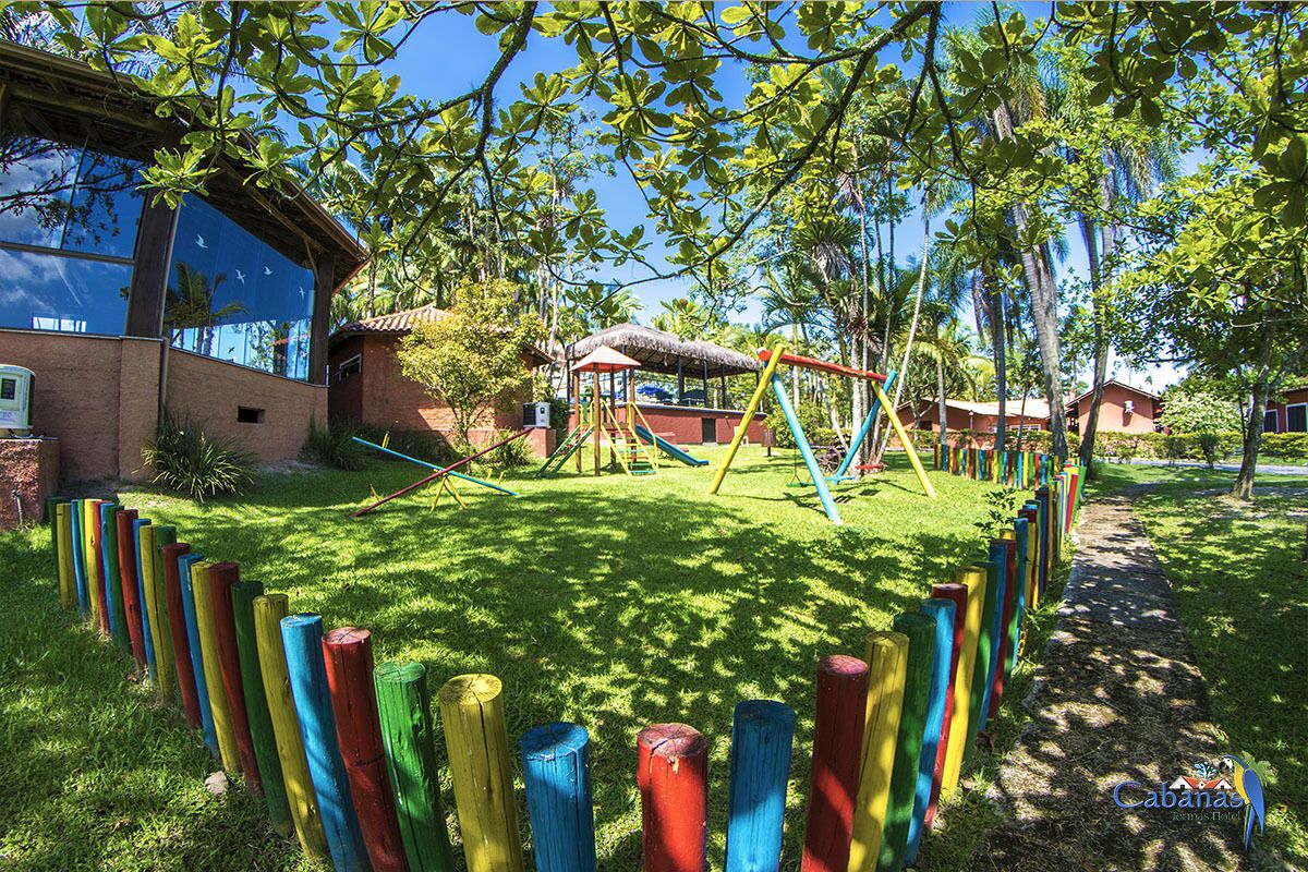 A playground with colorful wooden play structures and a thatched-roof shelter, surrounded by a grassy area and trees.