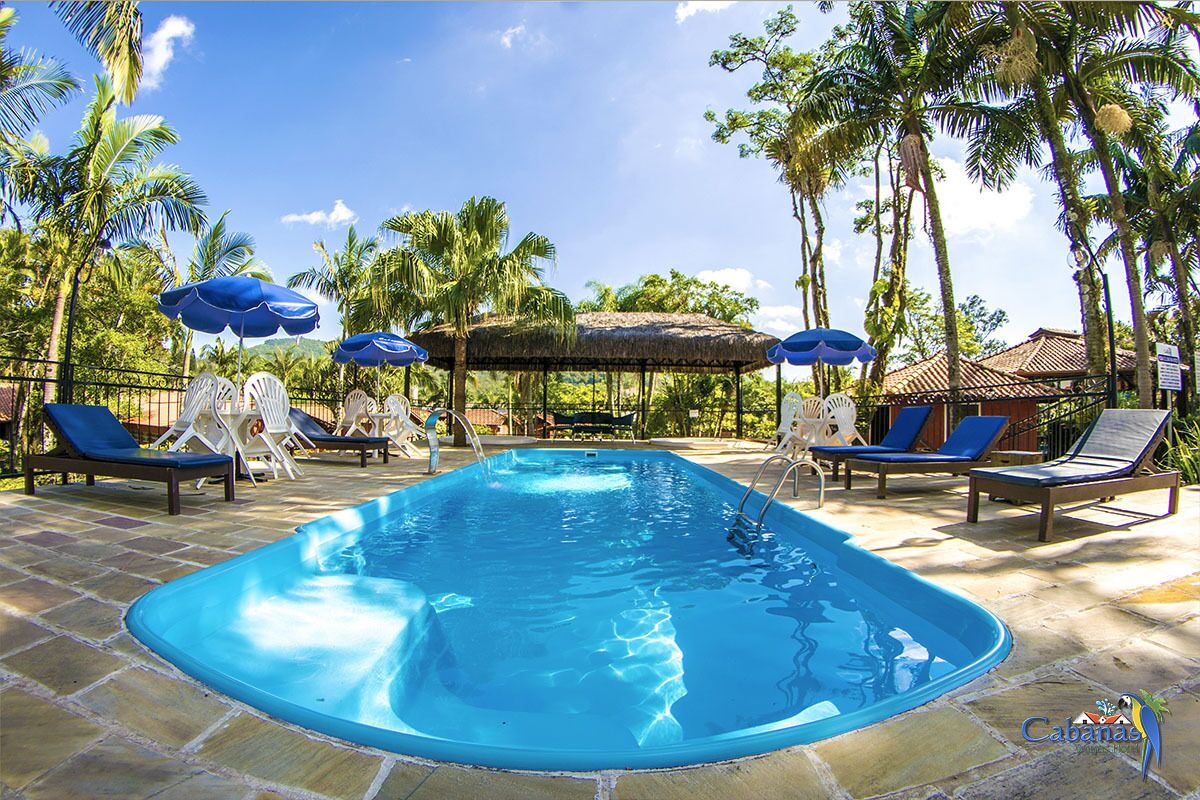 A rectangular swimming pool with lounge chairs and umbrellas, surrounded by palm trees and a thatched-roof structure.
