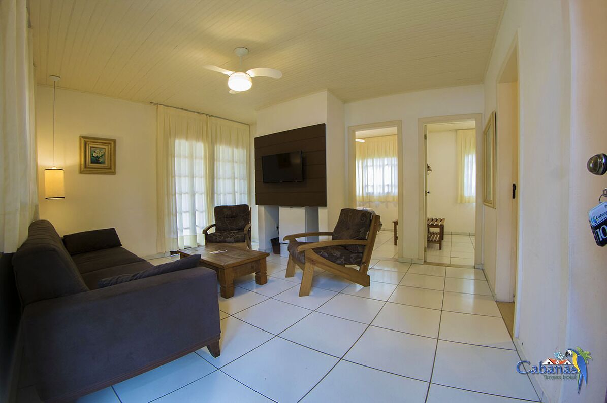 A living room with a sofa, two armchairs, a coffee table, a flat-screen TV mounted on the wall, and a ceiling fan.