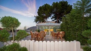 An outdoor seating area with wicker chairs and a glass enclosure, surrounded by a white picket fence and trees. - Hatunca Otel & Restaurant (Amasya)