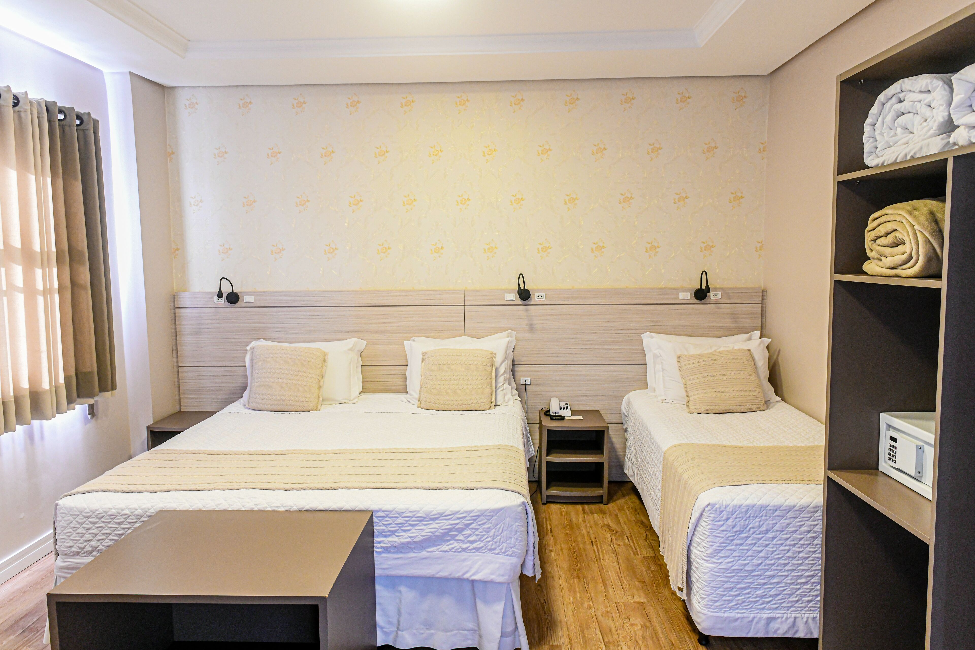 A hotel room with two beds, a bedside table, a shelf with towels, and a wall with a floral pattern.