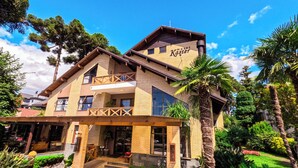 Et toetagers hotel med balkon, et skilt der siger "Hotel Kaster", og en velholdt have med palmer og buske.