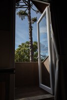 Puerta de balcón abierta con vista a un árbol alto y un cielo despejado.