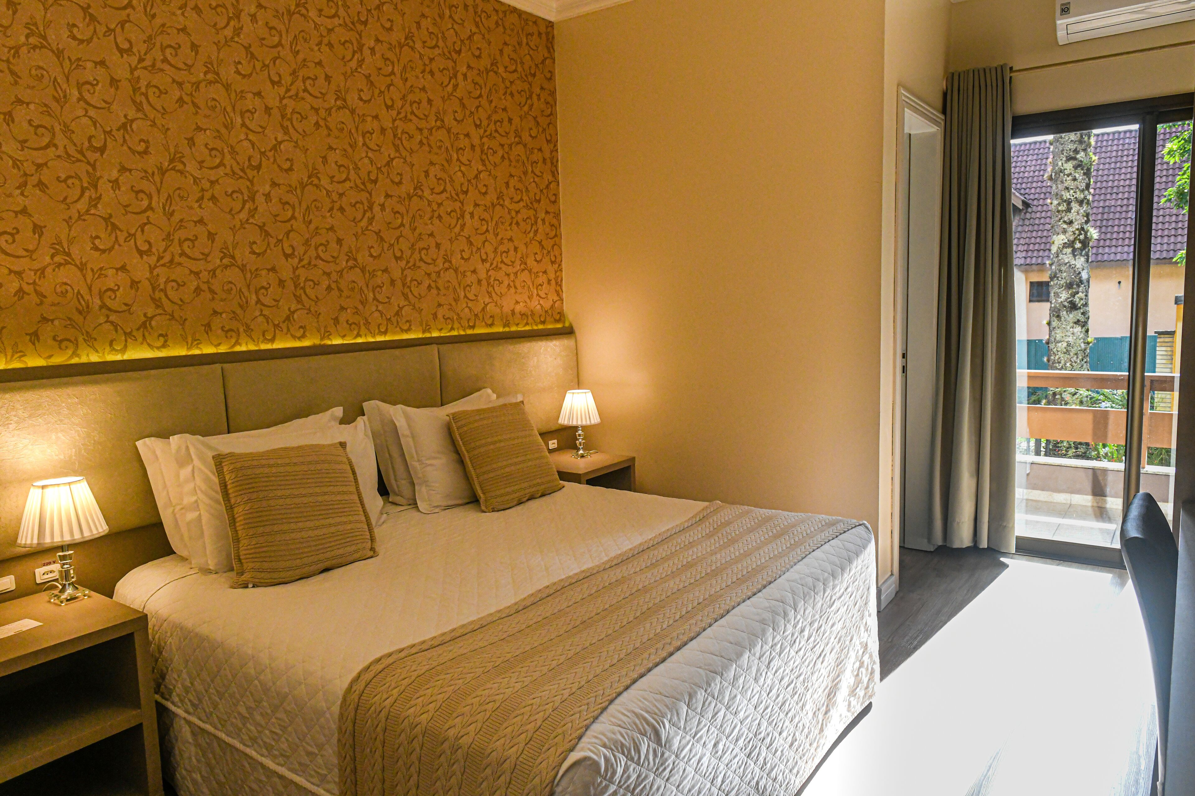 A hotel room with a large bed, two bedside lamps, a patterned wallpaper, and a view of the outside through a sliding door.