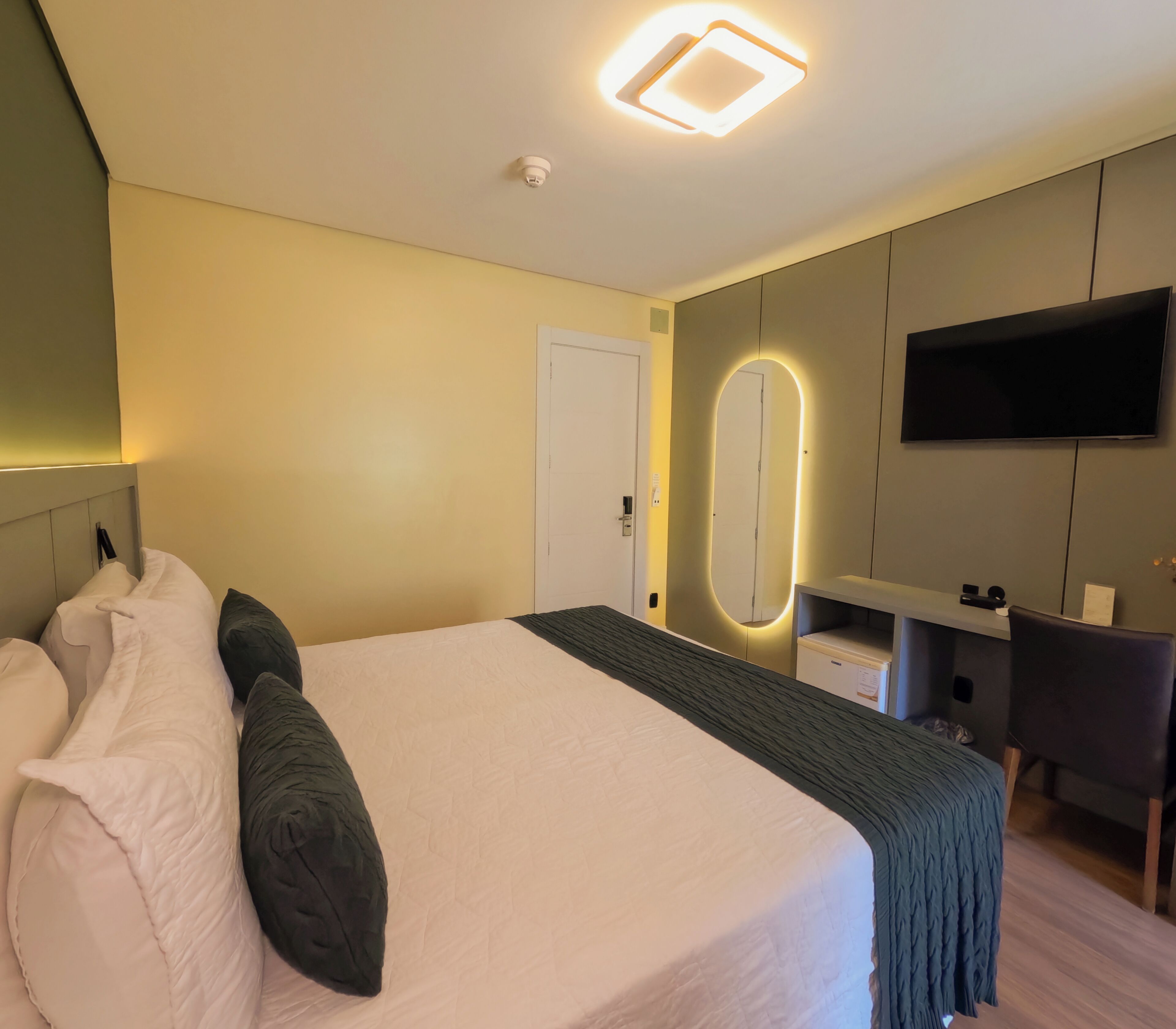 A modern hotel room with a large bed, a bedside table, a mirror, and a flat-screen TV mounted on the wall.