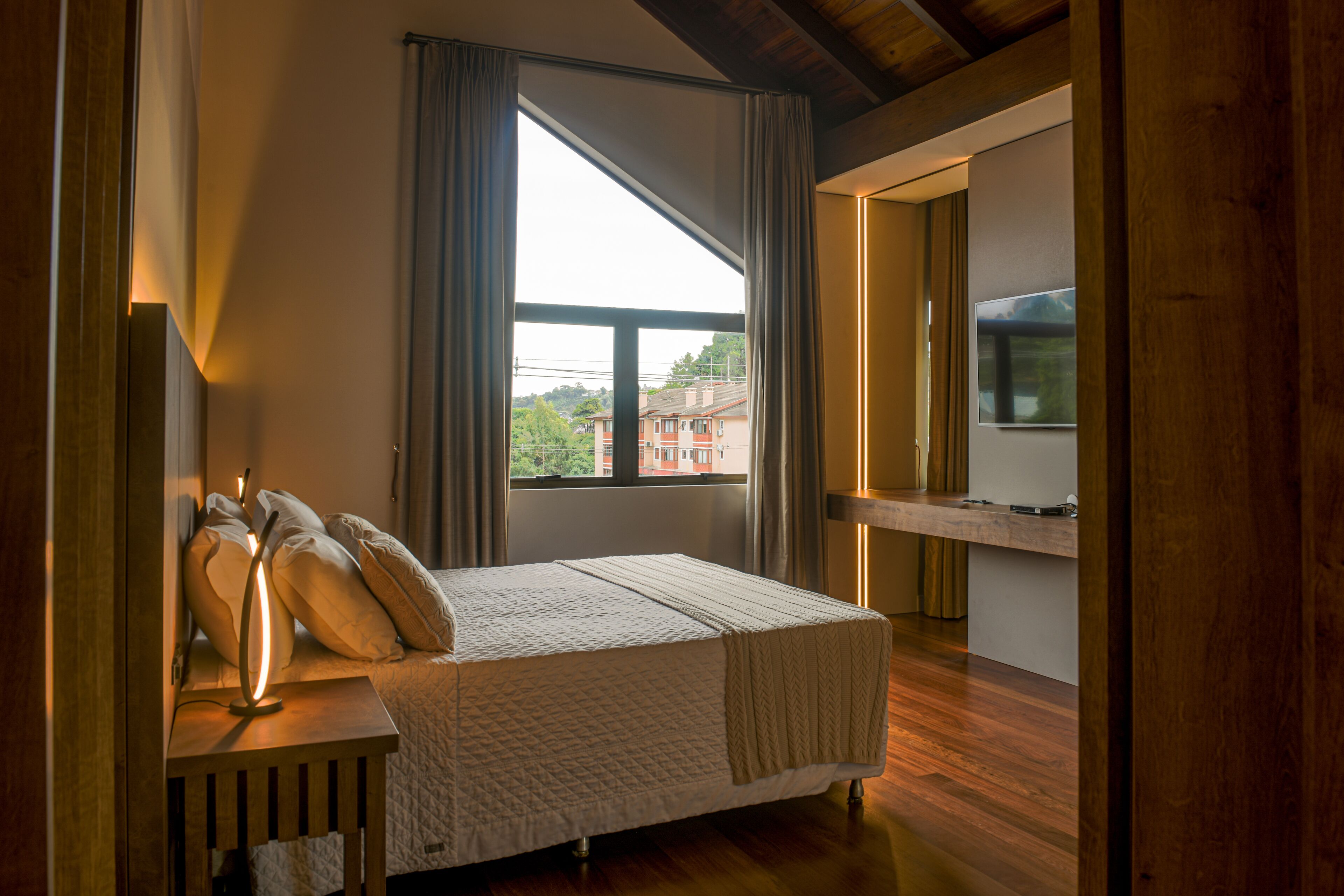 Una camera da letto con una grande finestra, un comodino con una lampada e un letto con cuscini e coperta.