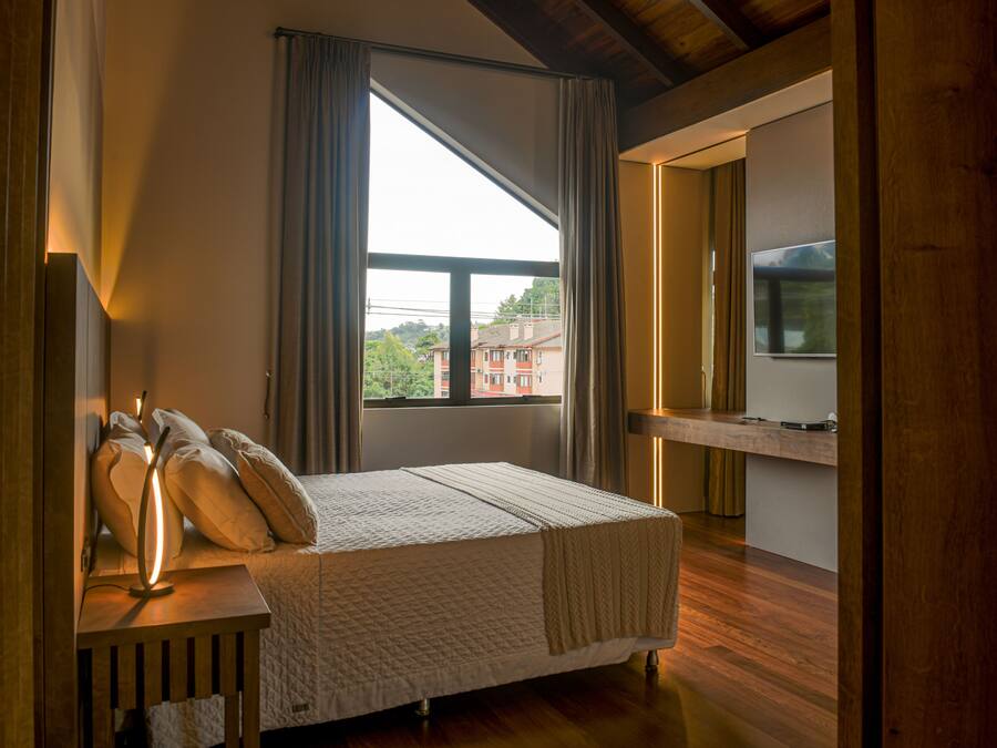 Una camera da letto con una grande finestra, un comodino con una lampada e un letto con cuscini e coperta.