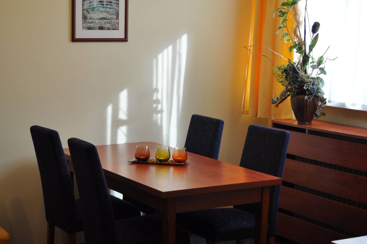 Ein Essbereich mit einem Holztisch, vier Stühlen, einer Kristallleuchtre, einem gerahmten Bild an der Wand und einem Blumentopf am Fenster.