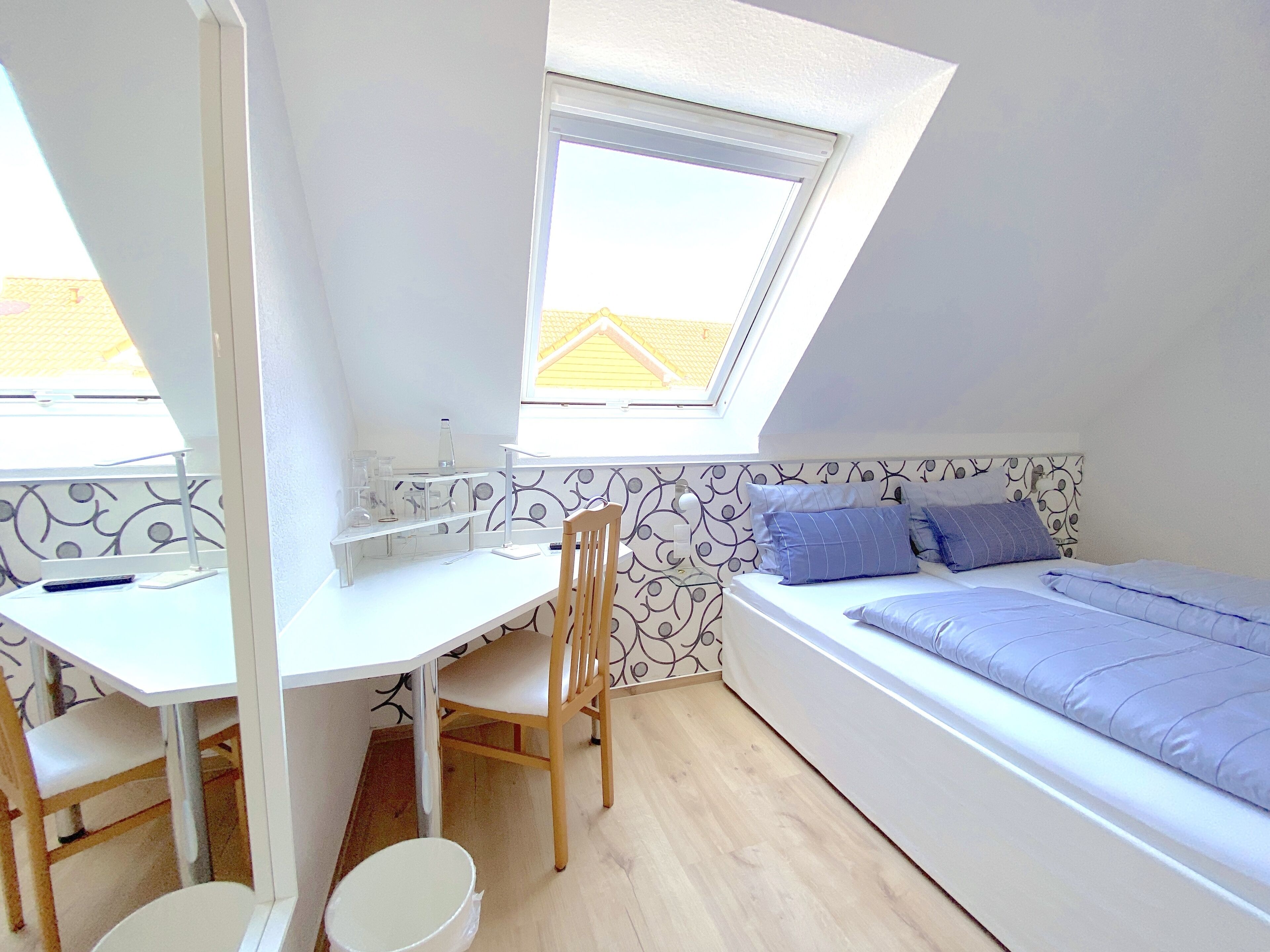 A compact room with a sloped ceiling, a desk with a chair, a bed with blue bedding, and a window with a view of a yellow building.