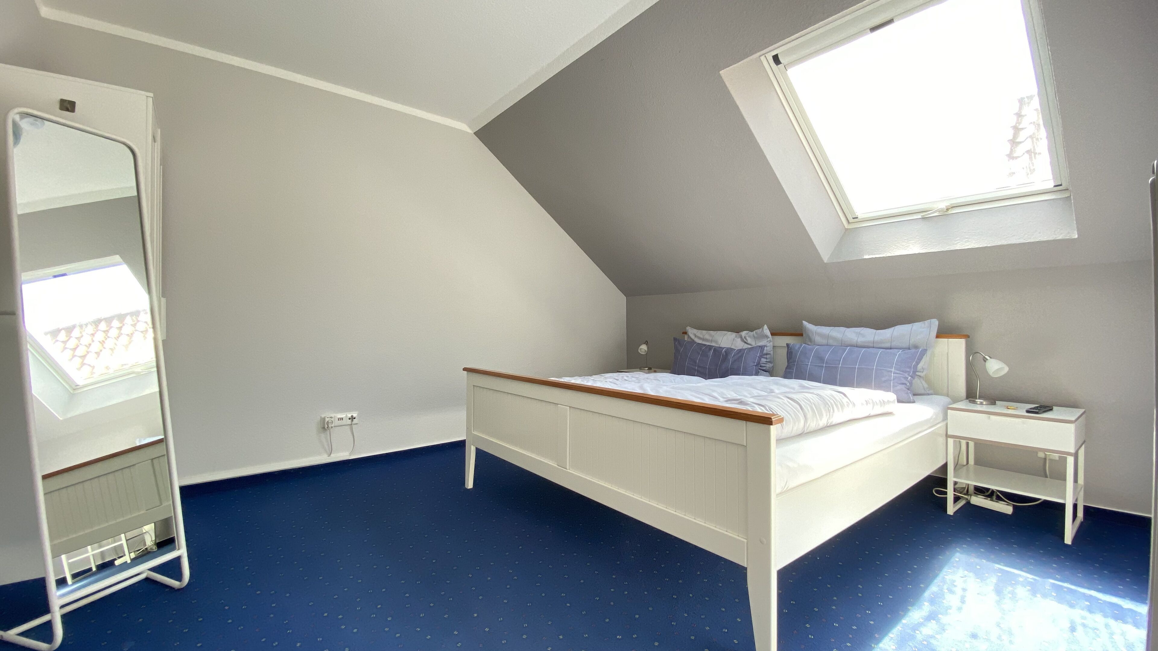 A bedroom with a sloped ceiling, a bed with white bedding, a bedside table with a lamp, and a large window providing natural light.