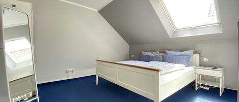 A bedroom with a sloped ceiling, a bed with white bedding, a bedside table with a lamp, and a large window providing natural light.