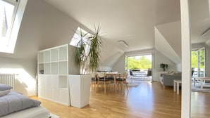 A spacious loft apartment with a high ceiling, wooden flooring, a dining area with a table and chairs, a gray sofa, and a large window providing natural light. - Hotel zur Bever (Telgte)
