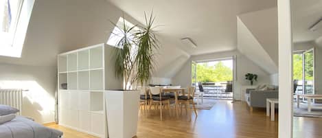 Un amplio apartamento estilo loft con techo alto, piso de madera, zona de comedor con mesa y sillas, sofá gris y un gran ventanal que proporciona luz natural.
